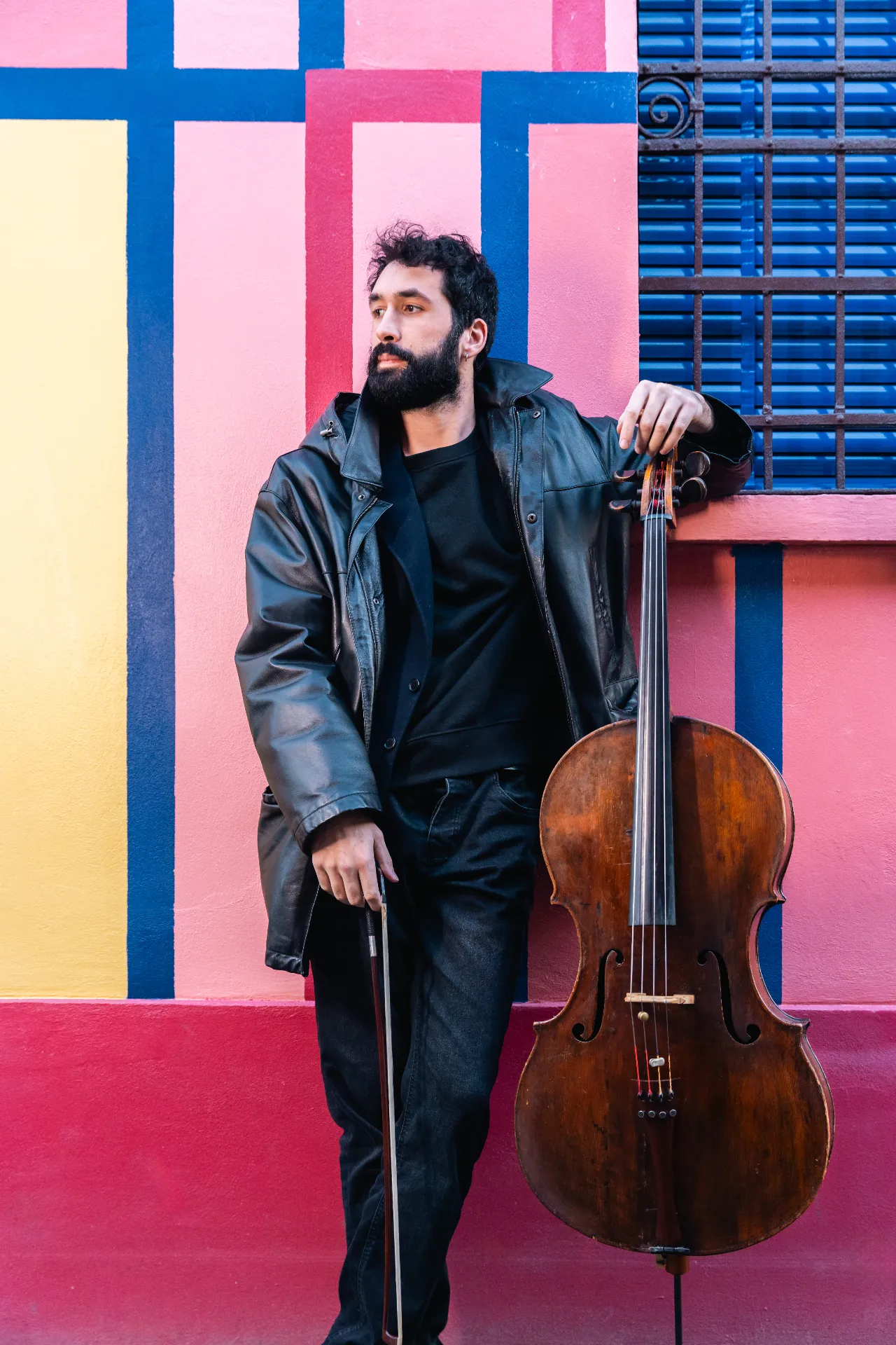 Portrait of Antonio Cortesi leaning against a colorful wall while holding the cello